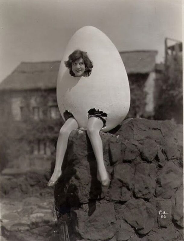 A woman sitting on a wall wearing an egg with a hole for her heads and holes for her legs.