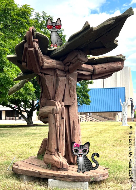 Periwinkle Stinky Charles Brouwer Sculpture of aa kneeling person with arms spread and with a cross/slash wings on its back with two cartoon tabby cats.