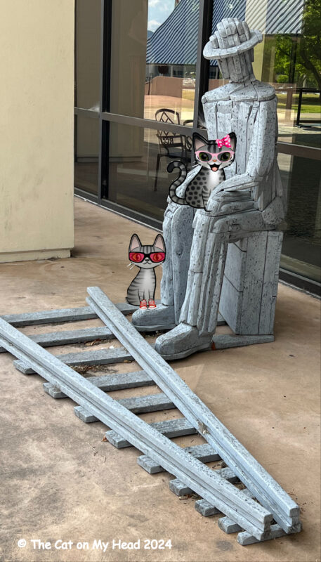 Periwinkle Stinky Charles Brouwer Tabby cat sitting in the lap of a sculpture of a man wearing a hat sitting next to a railroad track with another cartoon tabby cat sitting at his feet.