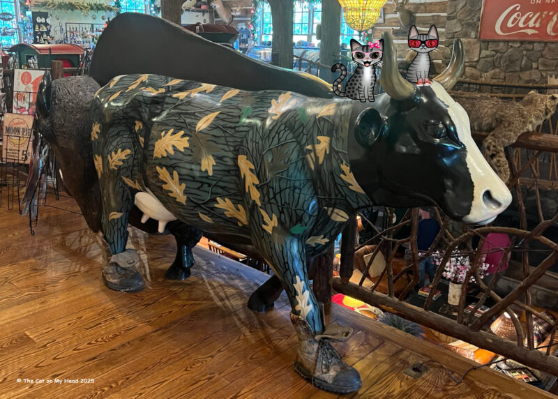 Two cartoon tabbies cats, Periwinkle & Stinky, riding on the statue of a cow wearing shoes at the Paint Bank General Store.